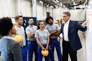 Mature businessman presenting business strategy on white board to group of industrial workers in a factory.