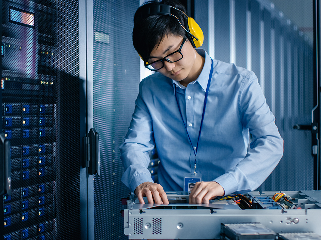 In Data Center: IT Engineer Wearing Protective Muffs Installs New Hardware for Server Rack. Specialist Doing Maintenance, Running Diagnostics and Updating Hardware for Stable Functioning of a System.