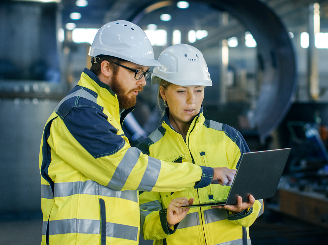 Two engineers wearing hi-vis jackets in a factory inspect a laptop screen