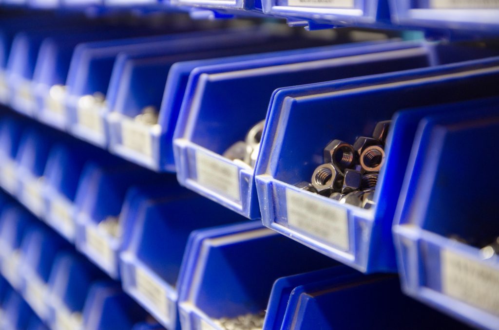 Rows of blue plastic storage bins containing stocks of parts and components, such as screws, bolts and nuts, in an industrial or commercial warehouse shot with selective focus